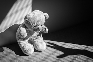 Black-and-white photo of a worn teddy bear sitting alone in striped window light, casting a long shadow on the floor; the scene feels quiet and melancholic.